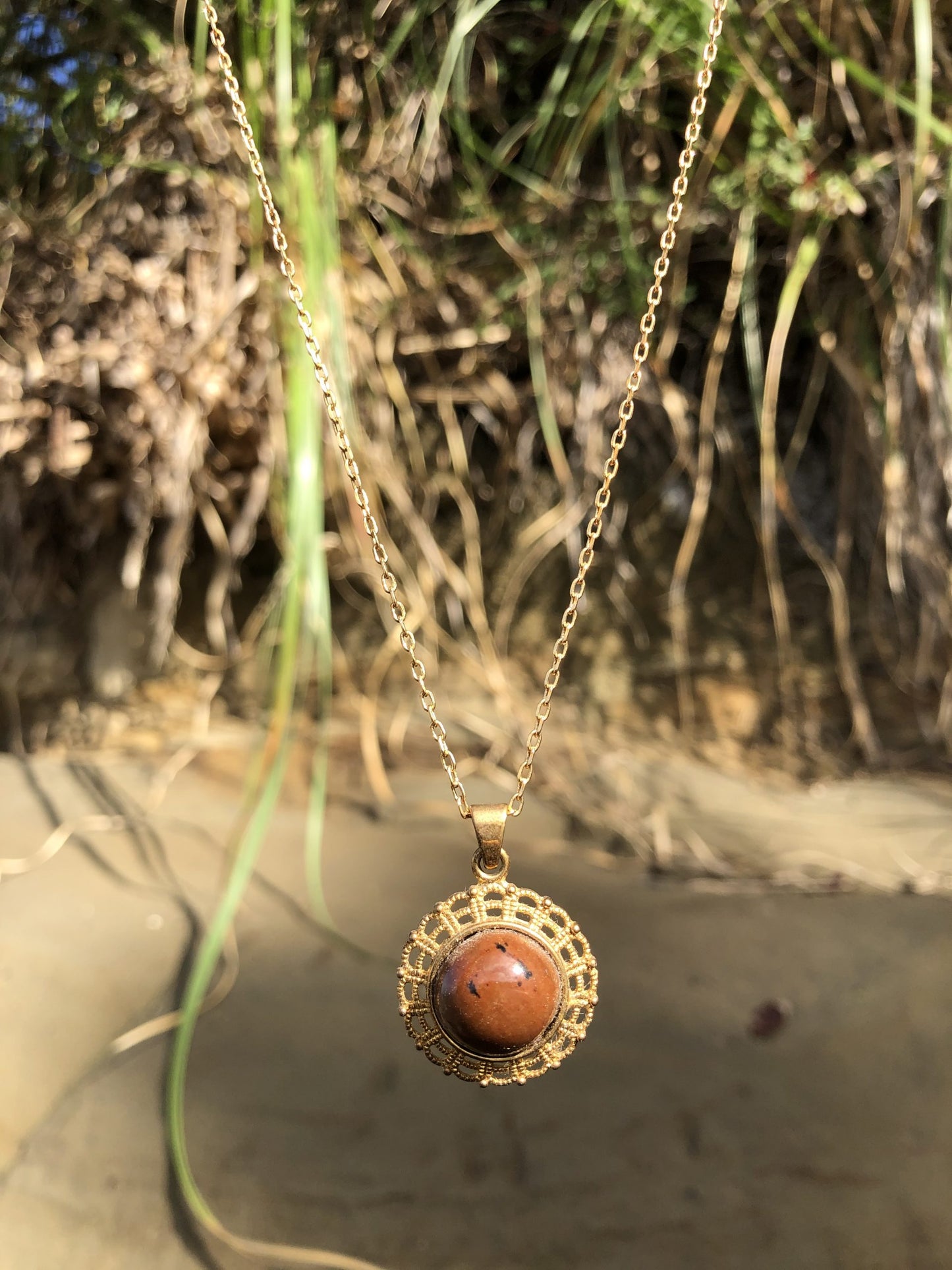 Necklace with New Zealand Mahogany obsidian, redish-brown with black spots, hand polished to a 14mm round cabochon and set in a gold plated setting with 19 inch chain, on beach
