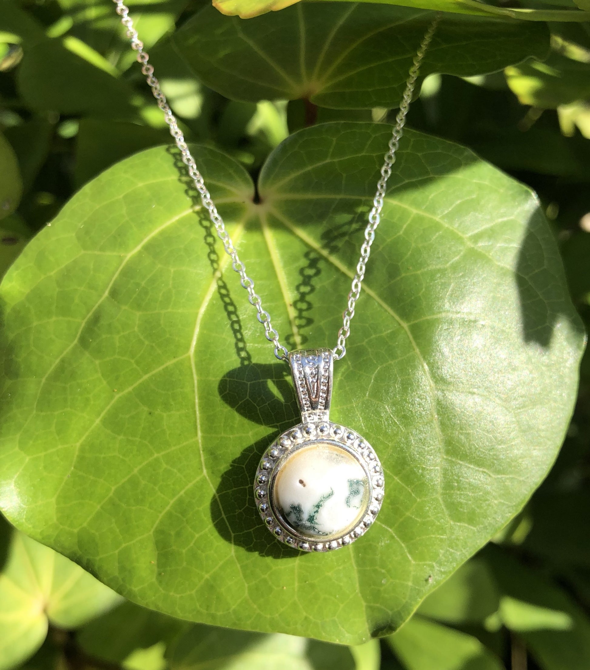 Necklace with natural Tree-Agate showing dark green moss on a bright white background, hand polished to a 10mm round cabochon and set in a silver plated setting with 19 inch chain, on leaf