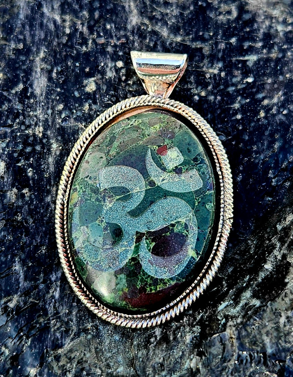 Necklace with a New Zealand multi-coloured rhyolite, hand polished to a 30x22mm cabochon with the Ohm symbol carved into the surface. This beautiful stone is set in a sterling silver setting with a 19 inch sterling silver chain, front.