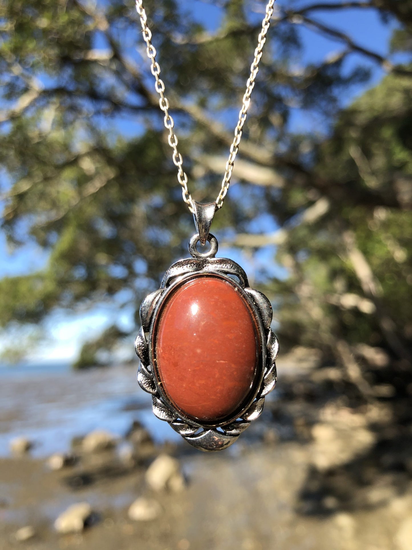 Necklace with New Zealand red Jasper, rich, deep red with tiny flecks of colour variation, hand polished to a 25x18mm cabochon and set in a silver plated setting with 19 inch chain, held up in front of tree on beach