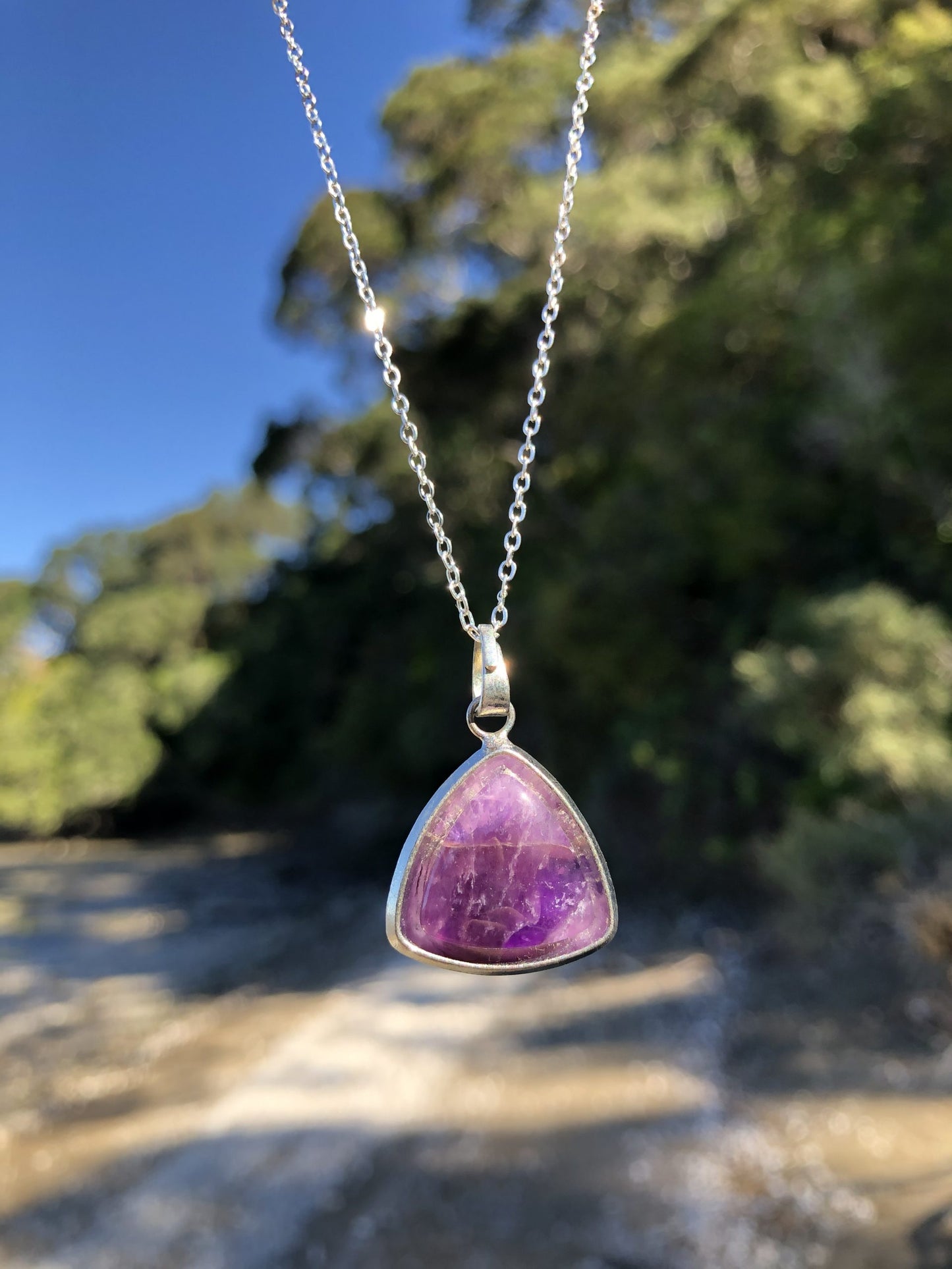 Necklace with rich, dark purple Amethyst, hand polished to a 18x18x18mm triangular cushion cabochon and set in a silver-plated setting with 19 inch chain on beach