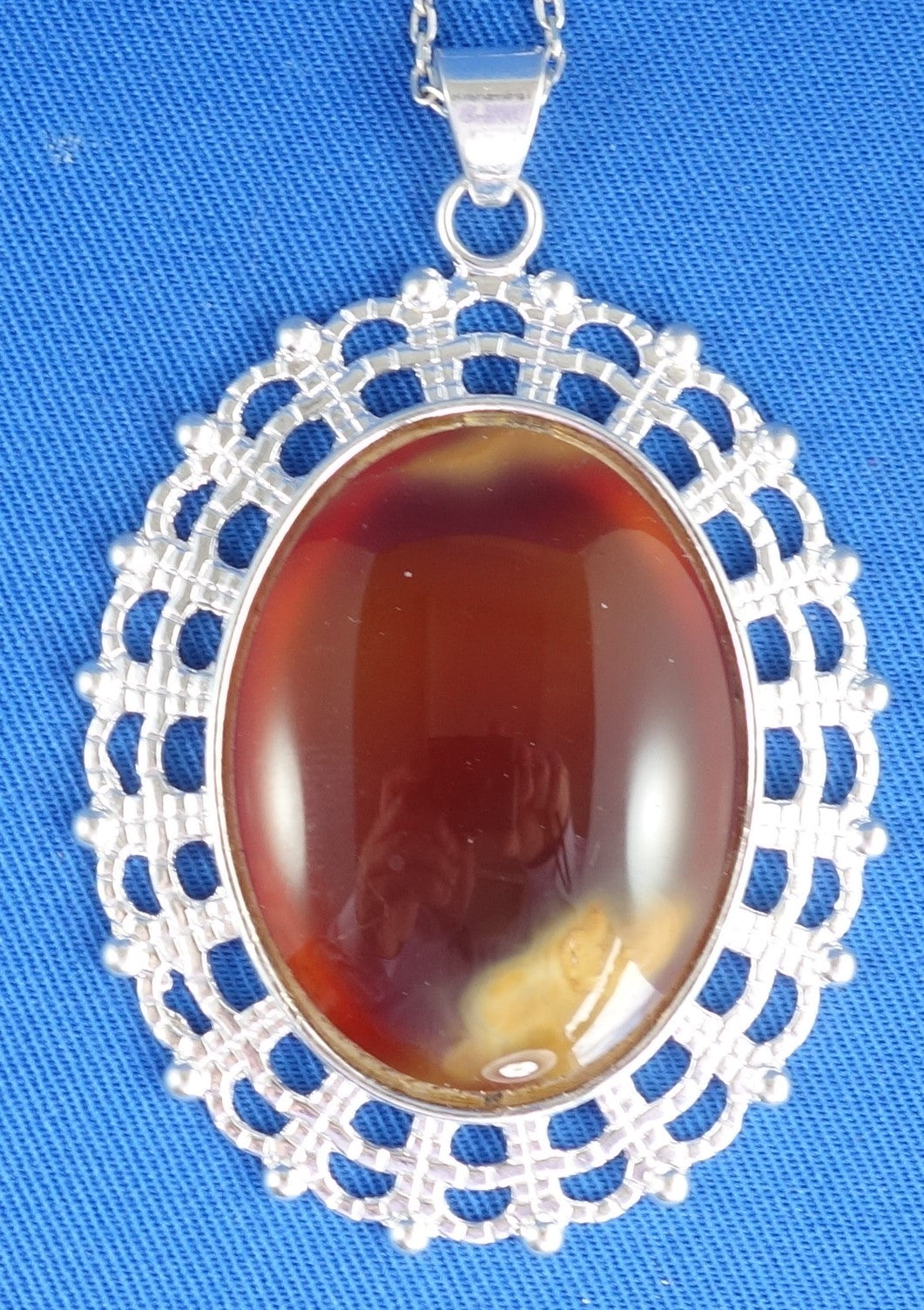 Necklace with large carnelian from Coromandel New Zealand with deep translucent orange color and side swirls of yellow/tan, hand polished to a 40x30mm cabochon and set in silver plated setting with 19 inch chain - on blue background