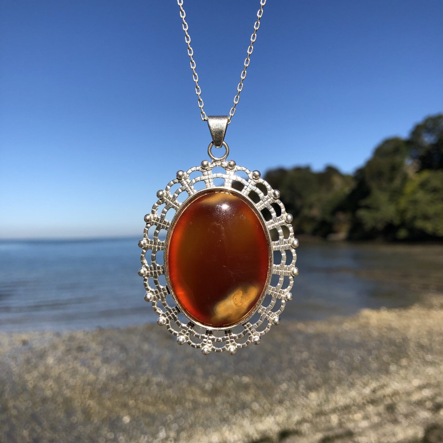 Necklace with large carnelian from Coromandel New Zealand with deep translucent orange color and side swirls of yellow/tan, hand polished to a 40x30mm cabochon and set in silver plated setting with 19 inch chain - on beach, front