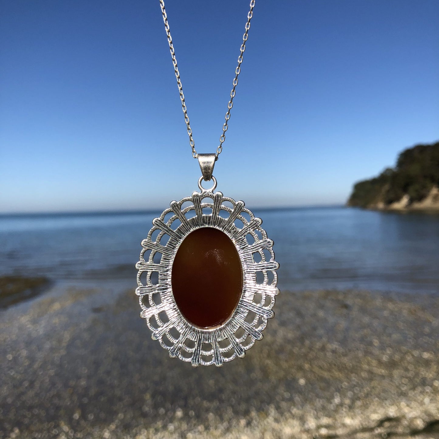 Necklace with large carnelian from Coromandel New Zealand with deep translucent orange color and side swirls of yellow/tan, hand polished to a 40x30mm cabochon and set in silver plated setting with 19 inch chain - on beach, back.