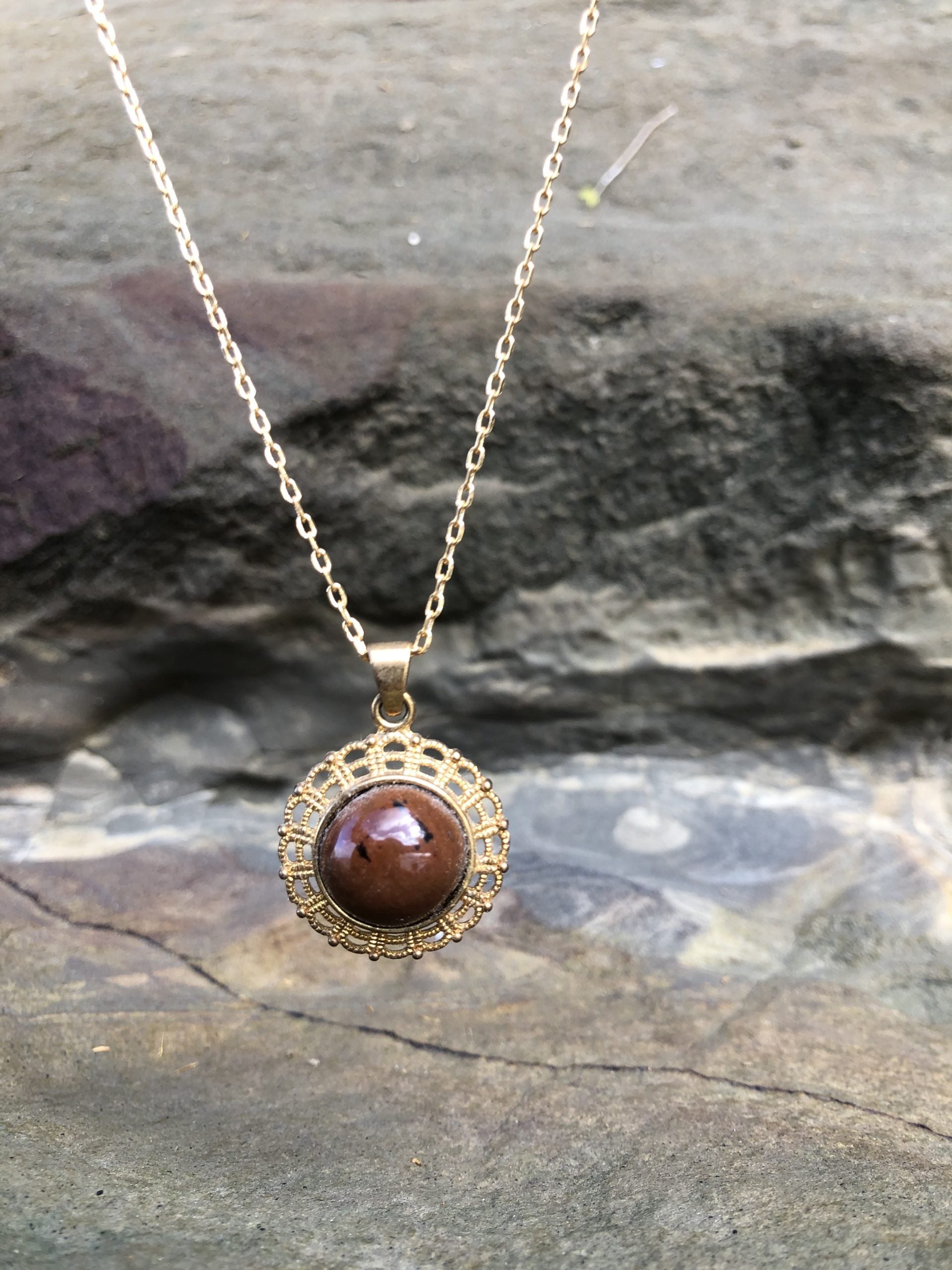 Necklace with New Zealand Mahogany obsidian, redish-brown with black spots, hand polished to a 14mm round cabochon and set in a gold plated setting with 19 inch chain, on rocks
