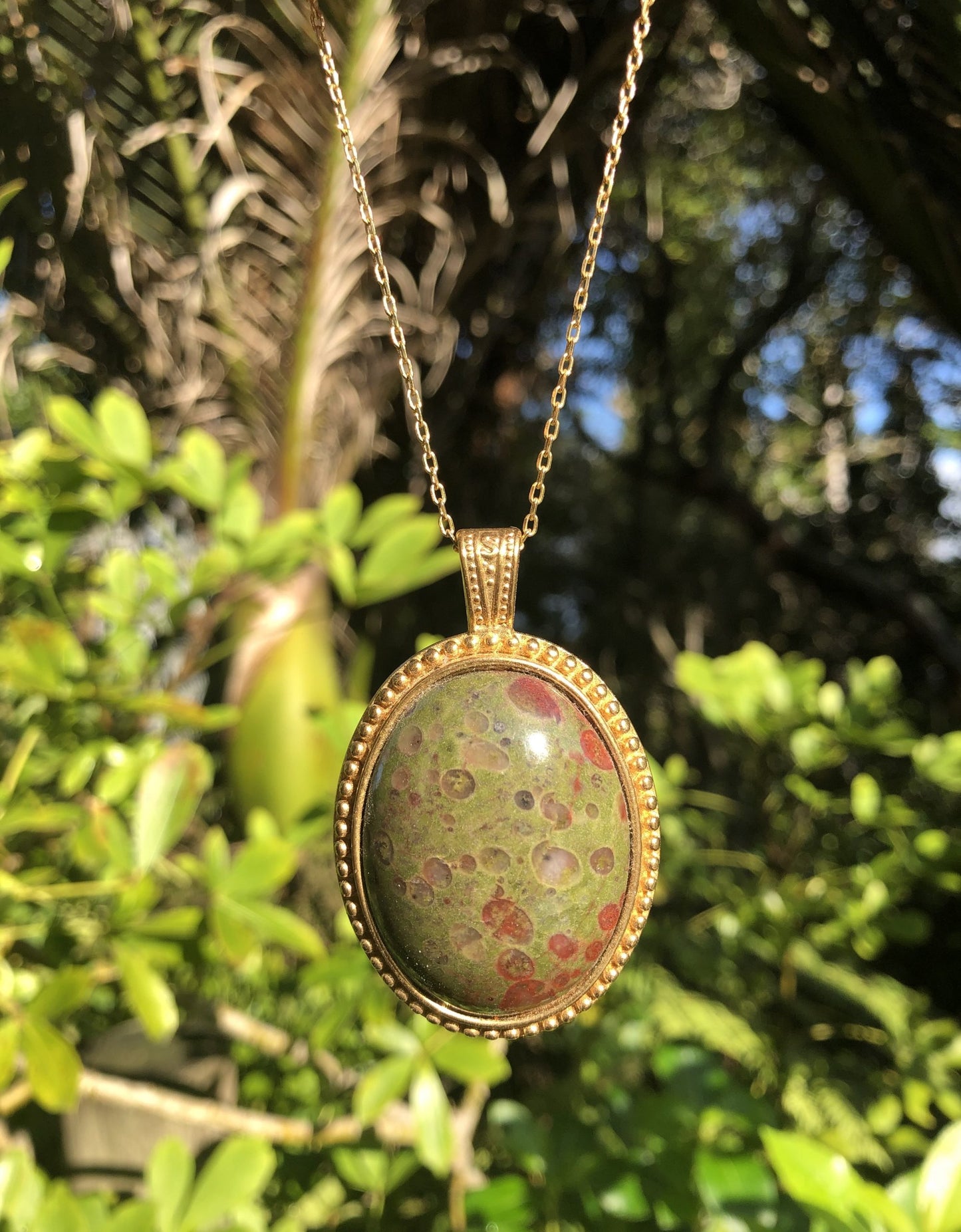 Necklace with rare New Zealand reverse poppy jasper with red poppy dots on a background of green jasper, hand polished into a 40x30mm cabochon and set into a gold plated setting with 19 inch chain - on leafy background